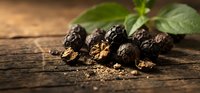 Whole black peppercorns with ground spice scattered on rustic wooden surface, fresh green leaves in background, warm natural lighting.