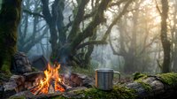 Campfire with metal mug in misty forest setting. Warm flames illuminate moss-covered rocks and ancient trees creating cozy outdoor adventure atmosphere.