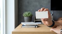 Person holding blank white business card in modern office workspace with wooden desk, notebook, and succulent plant.