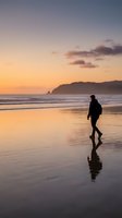 Silhouetted figure jogging along sandy beach at golden hour sunset, with calm ocean waves and distant headland, suitable for wellness and lifestyle content.