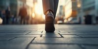 Close-up view of business person walking on urban sidewalk during golden sunset hour, showing leather shoe and suit pants against blurred city background.