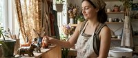 Young Caucasian woman artist in apron arranging ceramic animal figurines in a cozy pottery studio with natural light and plants.