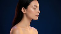 Young woman with closed eyes in profile view showcasing natural beauty and skincare routine against dark blue background for wellness and cosmetic concepts.