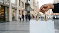 Person of Middle Eastern or South Asian descent holding blank white shopping bag on urban street with storefronts and pedestrians blurred in background for retail and commerce concepts.