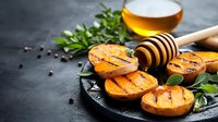 Grilled peach halves drizzled with honey on black slate plate garnished with fresh mint leaves and black peppercorns, golden honey dipper nearby.