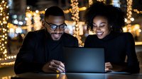 Young African American couple working on laptop at night, illuminated by warm holiday lights. Cozy winter atmosphere with bokeh background shows business or leisure time.