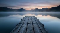 Wooden dock extending into misty lake at dawn with mountain silhouettes and pastel sky reflection creating peaceful morning atmosphere for meditation concepts.