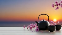 Traditional black teapot with cups and pink cherry blossoms on wooden table against sunset ocean view for zen meditation and relaxation concepts.