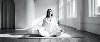 Young woman practicing meditation in white clothing sitting cross-legged on wooden floor in bright minimalist room with large windows for wellness.