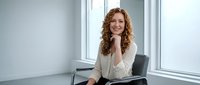 Professional businesswoman with curly red hair smiling confidently at modern office desk near window for corporate and workplace concepts.
