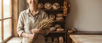 Happy male baker holding wheat stalks in artisan bakery with fresh bread display on wooden shelves creating warm authentic atmosphere for food business.
