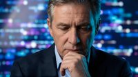 Middle-aged man with gray hair and facial stubble, wearing dark business suit and tie, hand thoughtfully positioned near chin, contemplative expression, set against blurred blue and purple digital technology background with bokeh lights, suitable for corporate, business, technology, and professional concepts.
