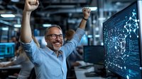 Excited mature Caucasian male programmer celebrating success at workplace, raising arms while looking at computer screen with data visualization.