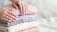 Woman hands touching soft knitted sweaters stack in pastel colors on wooden surface. Cozy winter clothing, textile care and laundry concept.