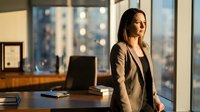 Young woman of South Asian descent, professional businesswoman wearing a tailored blazer, standing in modern office with floor-to-ceiling windows overlooking cityscape. Suitable for corporate, business, leadership, and professional development content.