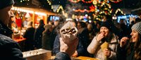 Young woman enjoying hot chocolate with marshmallows at festive Christmas market with twinkling lights and holiday decorations in background.