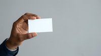 Man with dark skin tone holding blank white business card or paper against neutral gray background for corporate branding and identity mockup.
