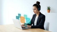 Young professional woman with headset working on laptop with digital real estate icons in modern office environment for remote property consultation.