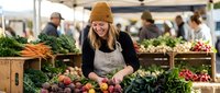 Young blonde woman in beanie hat selecting fresh vegetables at outdoor farmers market with organic produce displays and shoppers in background.