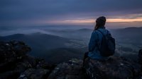 Hiker in blue jacket sitting on rocky mountain peak at dusk, gazing at misty valley landscape below during twilight hour for travel and adventure concepts.