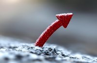 Red arrow pointing upward covered in frost and snow against blurred winter background, symbolizing growth, success and rising trends in cold conditions.