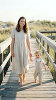 Smiling Caucasian mother in linen dress holds toddler son's hand on sunny wooden boardwalk near beach dunes, warm natural light.