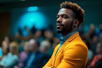 Confident African American businessman in bright orange suit jacket at conference, contemplative expression against blurred audience background.