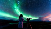 Woman in bathrobe observing northern lights through telescope against dramatic aurora borealis sky with stars and green glowing phenomenon over mountains.