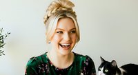 Young woman with blonde hair styled in an updo, fair complexion, wearing a dark green sequined top, smiling brightly while posing with a black and white cat for pet lover content.
