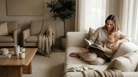 Young blonde Caucasian mother in beige sweater reading book while breastfeeding infant baby on neutral sofa in modern minimalist living room interior.