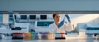 Female scientist in white lab coat examines test tube in modern laboratory surrounded by colorful sample vials, advancing medical research.