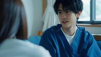Young Asian male medical professional in blue scrubs having conversation with patient in hospital room, warm natural lighting creates caring atmosphere.