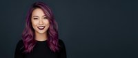 Young Asian woman with purple hair and dark lipstick smiling confidently against dark background for professional headshot and beauty concepts.