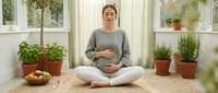 Young Caucasian pregnant woman meditating in lotus pose at home, hands on belly, surrounded by green plants, practicing mindfulness and prenatal yoga.