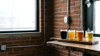 Craft beer flight with dark and golden ales in glasses on wooden board against exposed brick wall and industrial window.