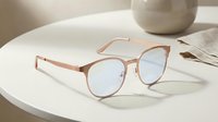 Rose gold round frame eyeglasses with blue light lenses on white table beside ceramic vase, suitable for fashion, eyewear, and lifestyle design projects.