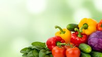 Fresh organic vegetables including bell peppers, tomatoes, cucumbers, cabbage and leafy greens arranged on soft green background for healthy cooking.