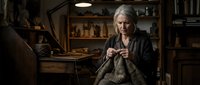 Mature woman with gray hair and fair complexion, wearing dark clothing, knitting in a rustic workshop filled with wooden furniture and vintage decor, warm lighting creating cozy atmosphere for crafts and hobbies.