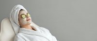 Woman relaxing in white bathrobe with cucumber eye mask and towel on head during spa treatment for skincare and wellness routine.