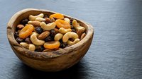 Mixed nuts and dried fruits in wooden bowl on dark slate surface for healthy snacking and cooking ingredients.