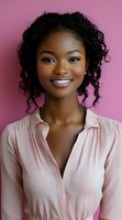 Young African American woman with natural curly hair and bright smile wearing pink blouse against pink background, professional headshot style portrait.