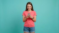 Young Caucasian woman in coral t-shirt using smartphone with happy expression on turquoise background for social media and technology concepts.