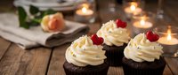 Chocolate cupcakes with vanilla frosting and cherry toppings on rustic wooden table with warm candlelight creating cozy bakery atmosphere for dessert photography.