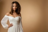 Mixed race woman in elegant white off-shoulder dress with flowing sleeves posing confidently against warm beige background for fashion and lifestyle content.