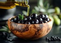 Fresh black olives in wooden olive bowl with pouring extra virgin olive oil, green leaves and dark moody background for food photography and design.