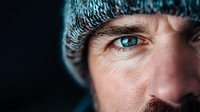Close up portrait of young Caucasian man with intense blue eyes wearing winter knit beanie hat against dark background, dramatic lighting highlights facial features.