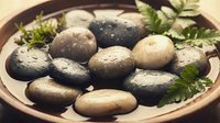 Fresh wet spa stones with water droplets and green herbs in wooden bowl for massage therapy and wellness relaxation treatments.