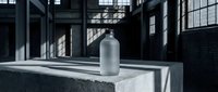 Frosted glass bottle on concrete surface in industrial warehouse with dramatic lighting and shadows through large windows for product mockup and branding.
