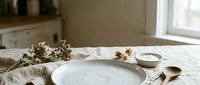 Minimalist table setting with white ceramic plate, wooden spoon, dried flowers, and neutral linen napkin on marble surface for dining and home decor styling.