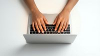 Hands typing on modern laptop computer keyboard from above view on white desk surface for business work and productivity.